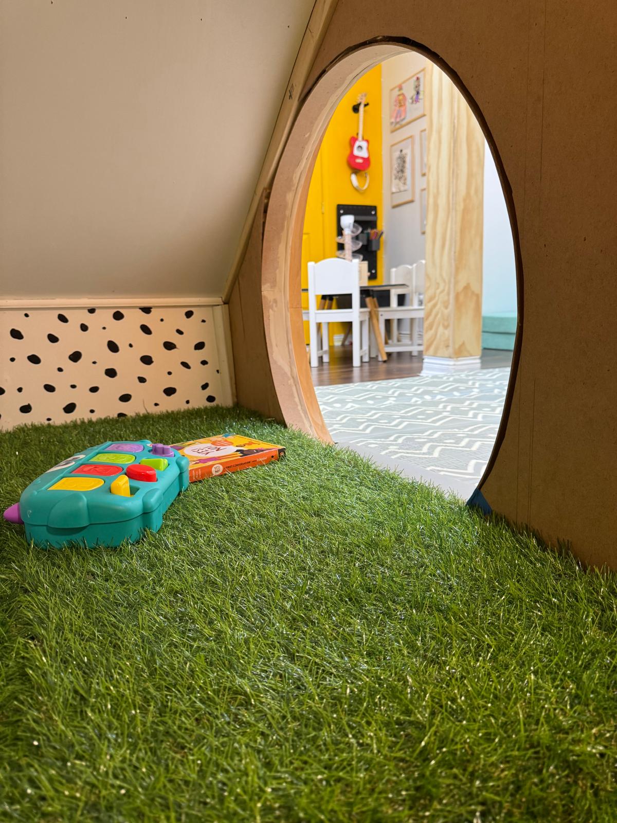 Looking out of a Funte den: an arched doorway cut through a plywood wall, artificial grass on the floor, a spotted skirting board, and the room beyond with a yellow wall, a toy guitar and a child's table.
