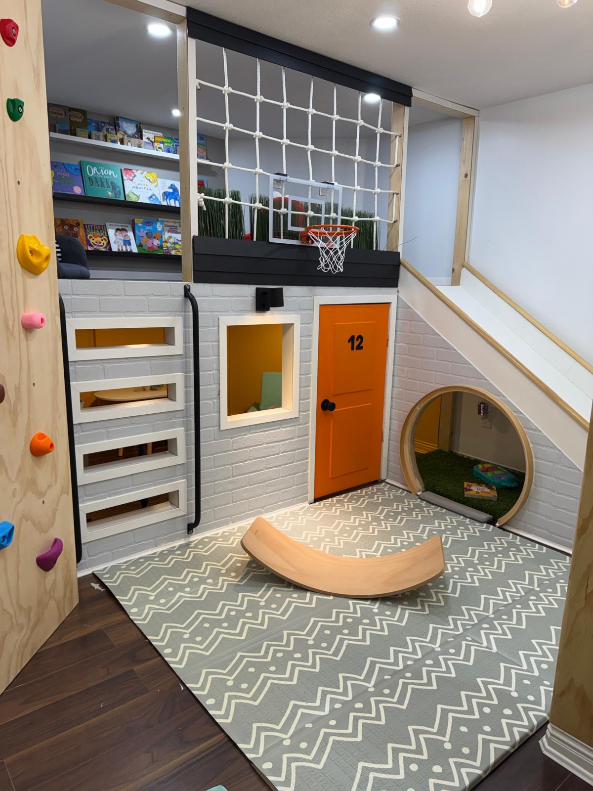 A finished Funte playhouse, photographed after it was built: a white brick front with an orange door numbered 12, a round tunnel opening beside it, a reading loft above behind a knotted rope net with a basketball hoop, a plywood climbing wall down one side and a wooden balance board on the rug.